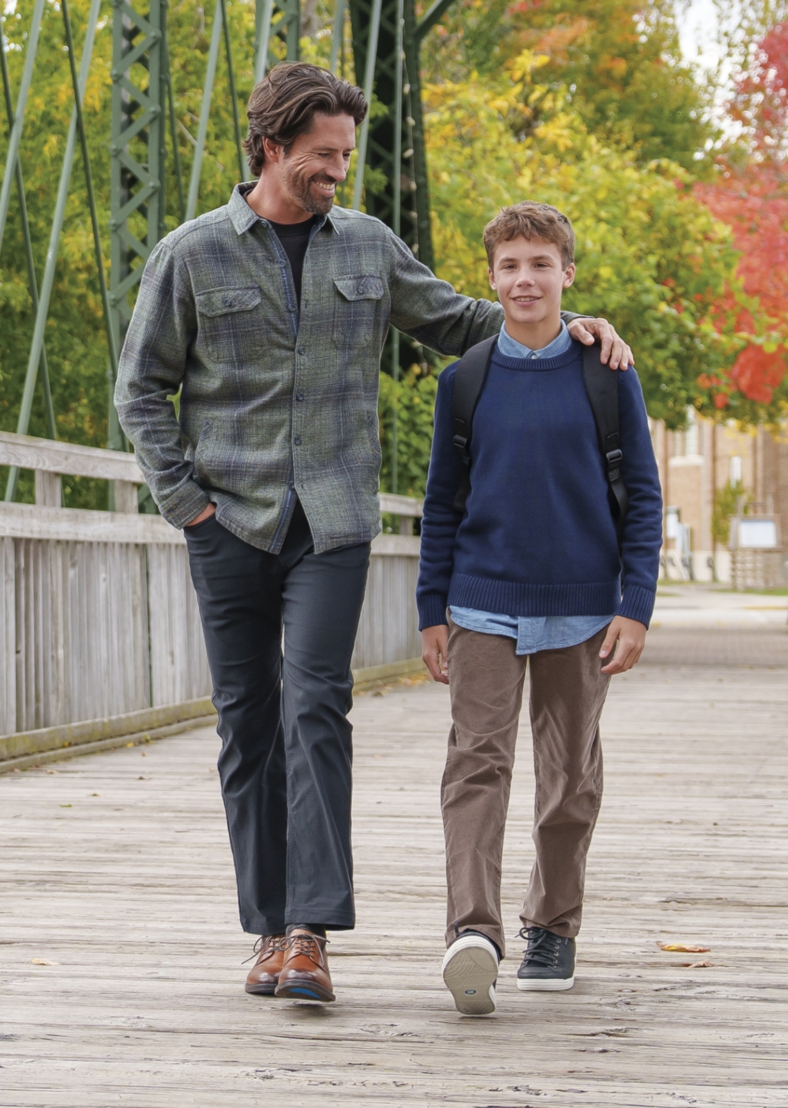 The featured image is a man wearing Nunn Bush cognac oxford dress shoes with his arm around his son while walking on a wooden bridge with fall foliage in the background.